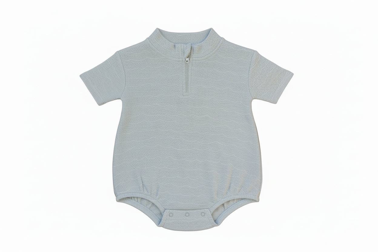 Light blue baby onesie with a zipper on a beige background with sandals and a plant leaf.