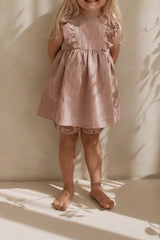 Young girl in a pink dress standing against a white curtain with sunlight casting shadows.
