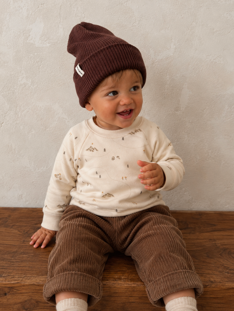 A baby sitting on a wooden floor wearing a dark brown ribbed knit beanie, a cream long-sleeve top with a small print, and brown corduroy pants against a textured light gray wall.