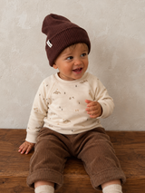 A baby sitting on a wooden floor wearing a dark brown ribbed knit beanie, a cream long-sleeve top with a small print, and brown corduroy pants against a textured light gray wall.