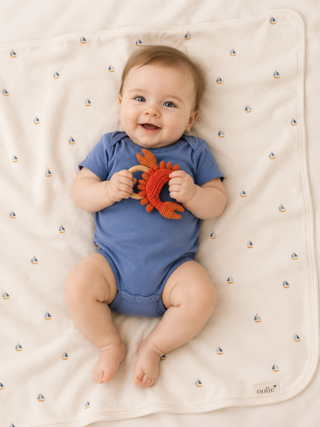 Baby in a blue onesie holding a red toy on a white blanket with sailboat pattern