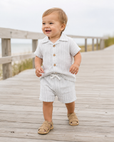 Baby in striped outfit standing on a wooden boardwalk near the beach