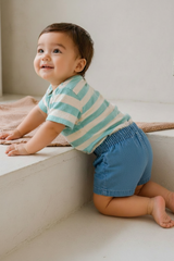 Child wearing a striped shirt and blue shorts sitting on a light-colored floor.