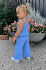 Child wearing a blue crochet outfit standing outdoors with plants and flowers in the background