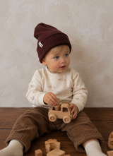 A baby sitting on a wooden floor wearing a dark brown ribbed knit beanie with a small white embroidered logo, a cream long-sleeve shirt, and brown corduroy pants, holding a wooden toy car.