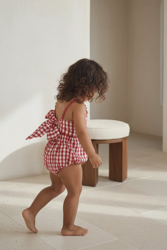 Child in a red and white checkered outfit standing near wooden chairs.