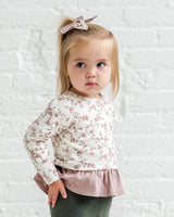 Young girl wearing a floral blouse and green pants against a textured white background
