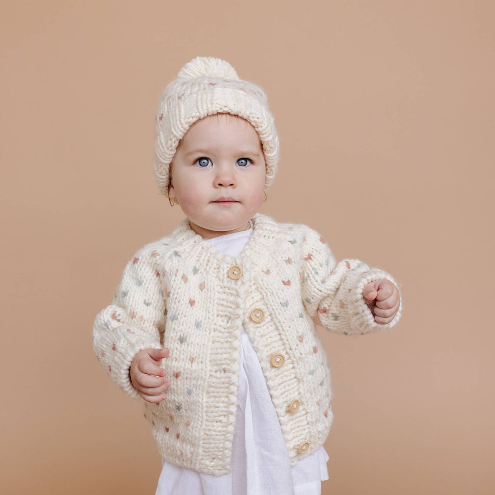 Baby wearing a cream-colored knitted cardigan and hat against a beige background