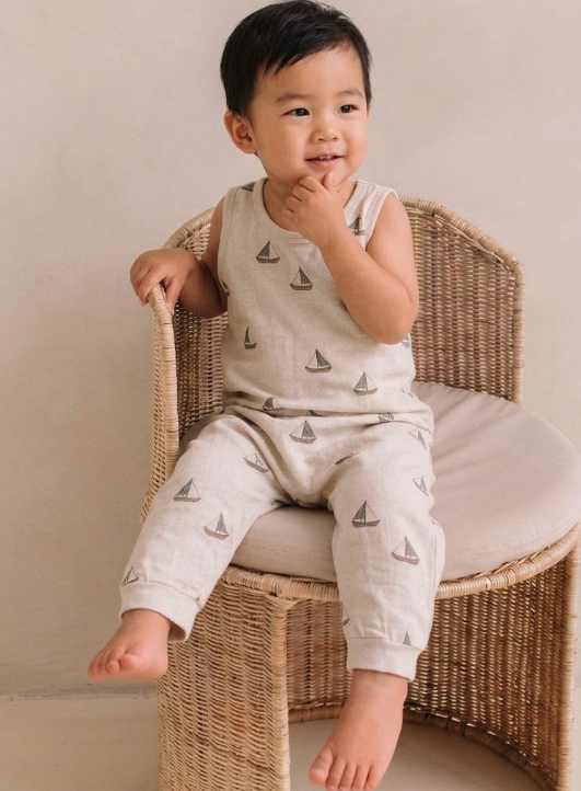 Child wearing a light gray sleeveless romper with sailboat pattern, sitting on a wicker chair.