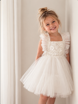 A young girl with blonde hair in a messy bun wears a white tulle flower girl dress with ruffled sleeves and a beaded bodice, standing against a neutral cream-colored wall.