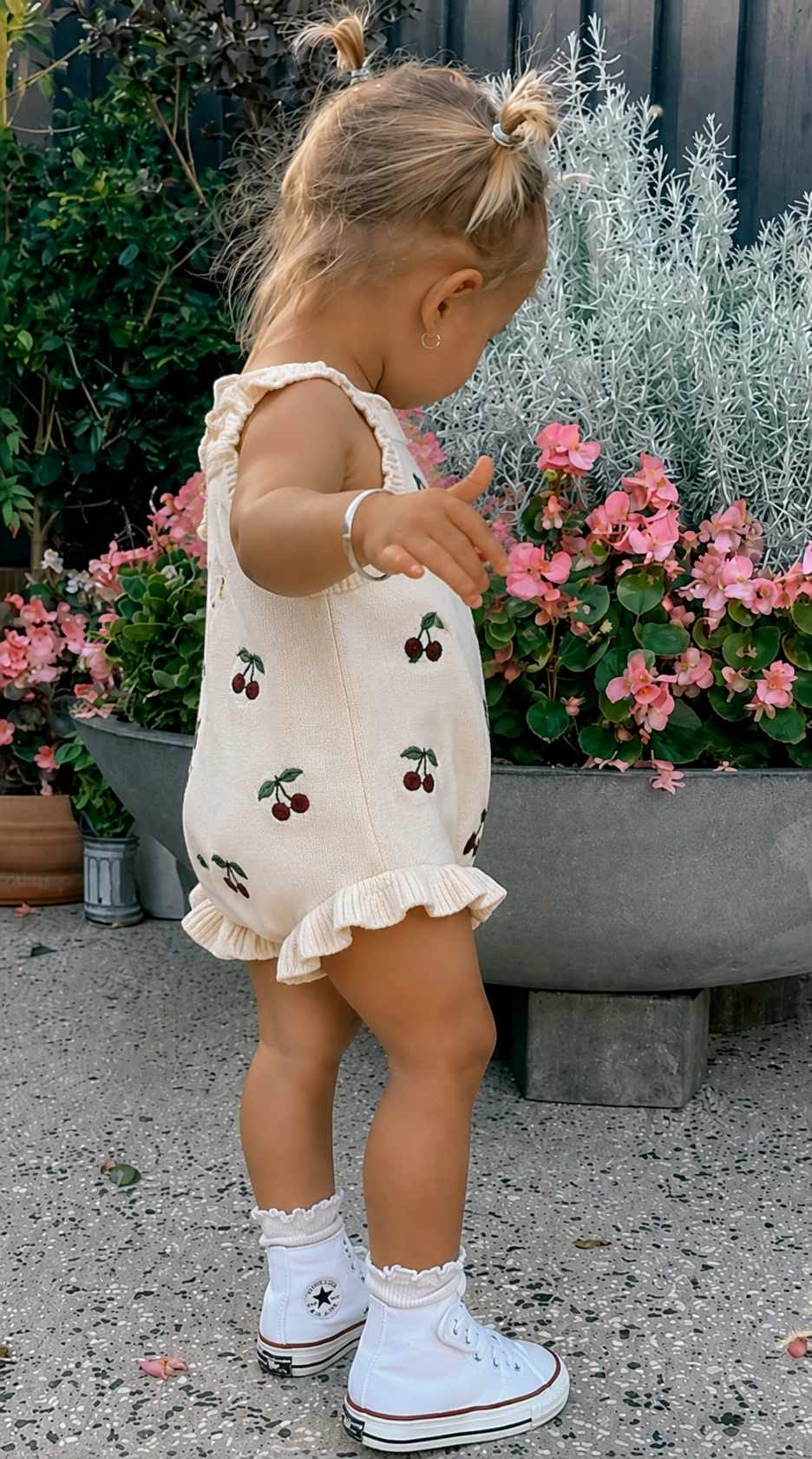 A toddler in a cream cherry-knit romper and high-tops reaching toward a bed of pink flowers.