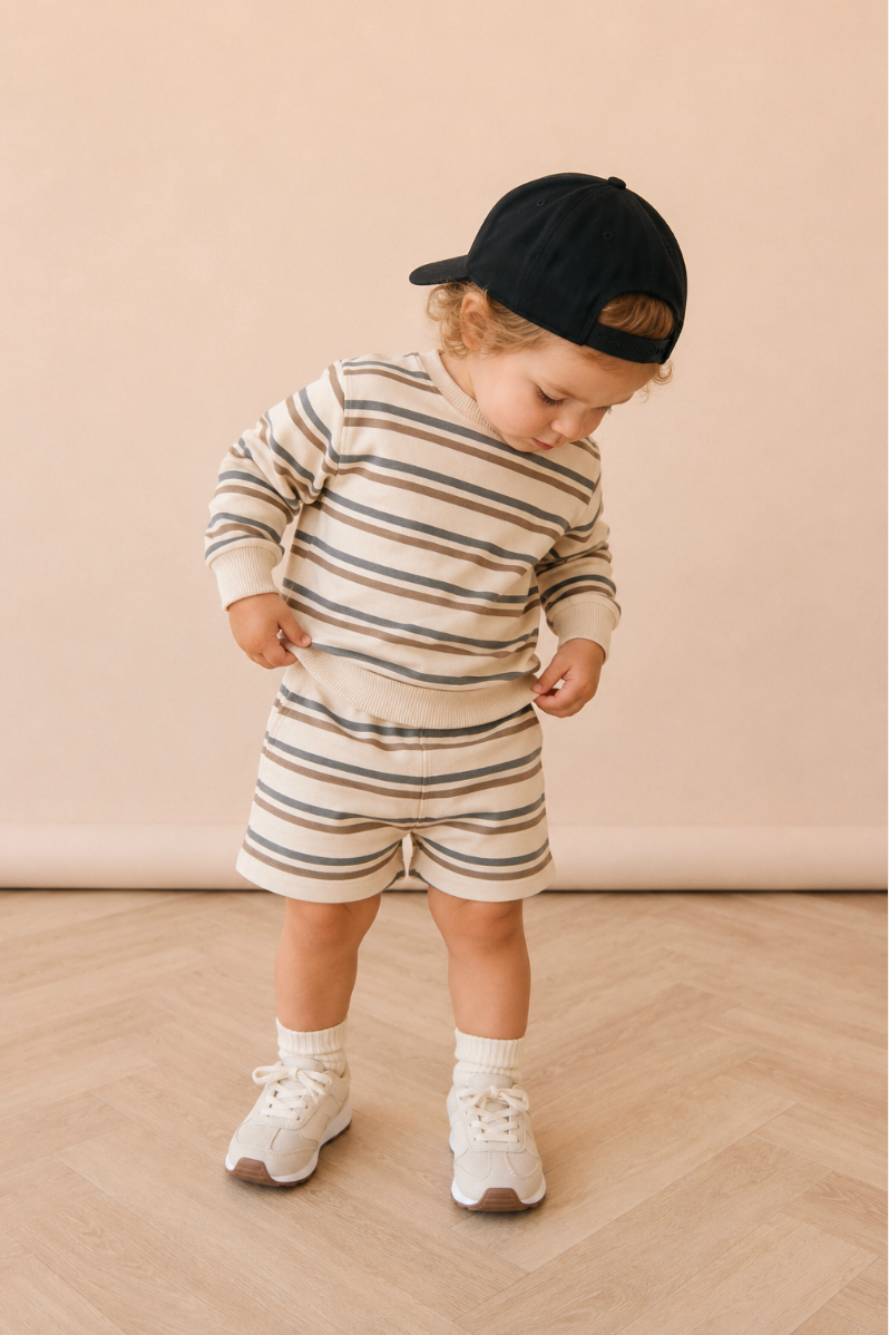 A toddler in a striped organic-cotton jumper set and cream sneakers, standing on a herringbone floor.